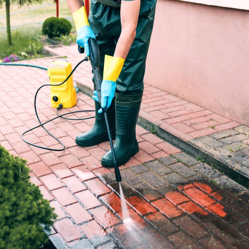 Man cleaning red concrete pavement block using high pressure water cleaner. Paving cleaning concept. Man wearing waders, rubber boots, gloves and waterproof trousers doing spring jobs in the garden