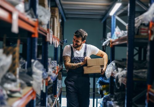 Hardworking smiling tattooed bearded worker relocating boxes while walking in storage of import and export firm.